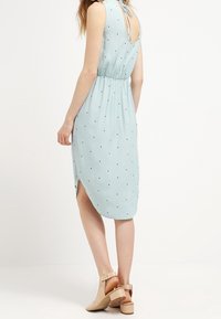 Vaaleansininen ilmanhihoja mekko, jossa on mustia pilkkuja, vyötäröllä on nyöri ja alaosassa halkio. Takana on nauhatuki.