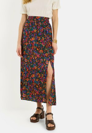 Mujer que lleva una falda midi floral y colorida con una apertura en el muslo, camiseta blanca de manga corta, pulsera dorada y sandalias marrones de plataforma.