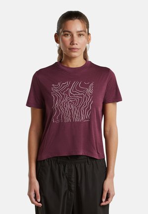 Frau mit braunen Haaren, die ein bordeauxrotes T-Shirt mit weißem abstraktem wellenförmigem Muster und schwarze Hosen trägt, steht vor einem schlichten Hintergrund.