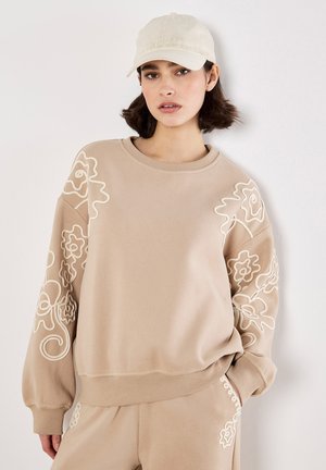 Beige Sweatshirt mit floralem weißen Stickmuster an den Ärmeln, geripptem Halsausschnitt und Bündchen. Getragen mit einem passenden Outfit und heller Cap.