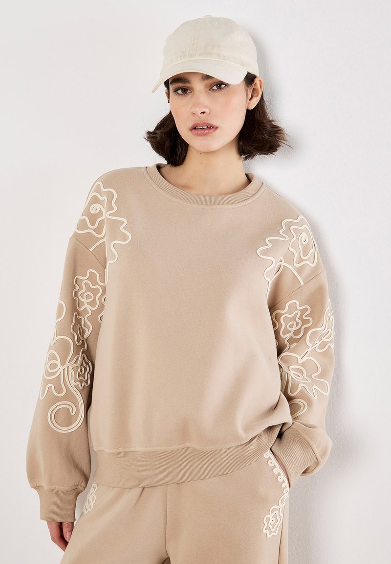 Sudadera beige con bordado floral blanco en las mangas, cuello y puños acanalados. Combinada con un atuendo a juego y una gorra de color claro.