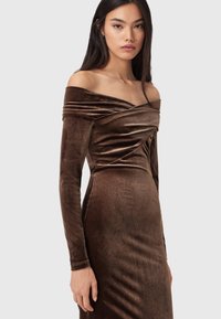 Brązowa aksamitna sukienka z odkrytymi ramionami i długimi rękawami, z drapowanym dekoltem i dopasowaną sylwetką, podkreślająca gładką teksturę.