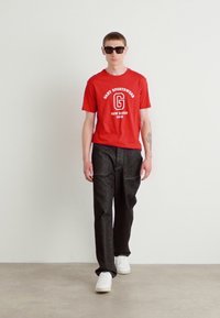 Rød bomulds T-shirt med hvid grafik og tekst, parret med mørke denimbukser og hvide sneakers. Solbriller fuldender outfittet.
