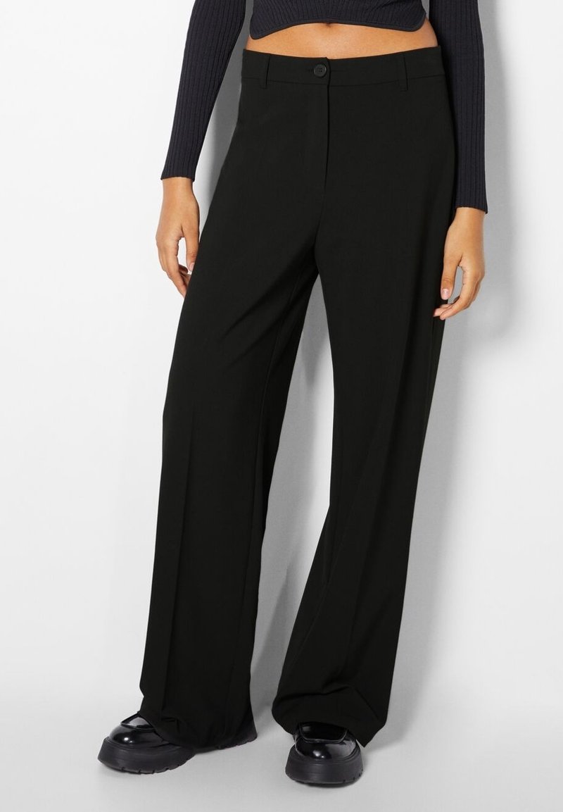 Zwarte, wijde broek gemaakt van gladde stof, met een knoopsluiting bij de tailleband en een strakke uitstraling. Gepaarde met chunky zwarte schoenen.