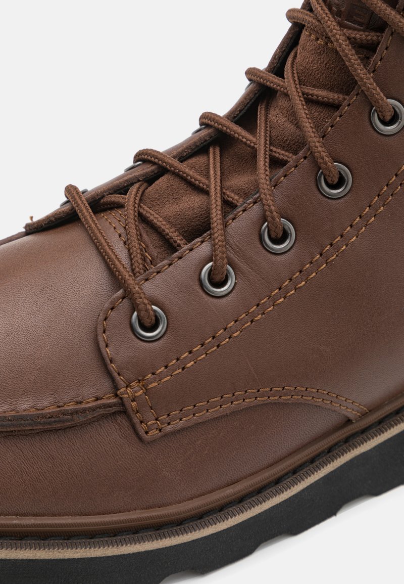 Botte en cuir marron avec œillets en métal, couture contrastante et lacets texturés. La semelle est noire, ajoutant durabilité et adhérence.