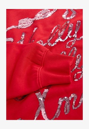 Sweatshirt rouge en tissu texturé, orné de paillettes argentées brillantes formant des motifs ondulés. Poignets côtelés avec une coupe ajustée.