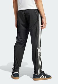 Svarta träningbyxor med vita randiga sidodekorationer, tryckknappsstängningar och ett varumärkeslogotyp vid knäet. Tillverkade av ett slitstarkt, lätt material.