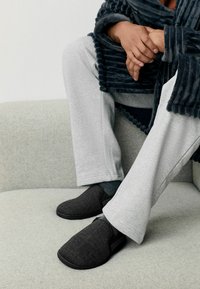 Încălțăminte din material textil negru, cu un design texturat, având un stil slip-on și talpa din cauciuc. Asortate cu pantaloni de trening gri pe o canapea deschisă.