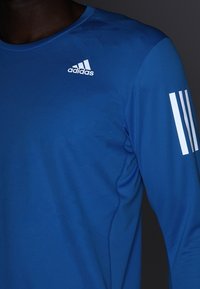 Athletické tričko s dlouhým rukávem v živě modré barvě, vyrobené z lehké látky. Na hrudi je bílý logo Adidas a na rukávu tři pruhy.