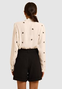 Camicia bianca a maniche lunghe con piccole ricami floreali, abbinata a pantaloni corti neri a vita alta con orlo risvoltato.