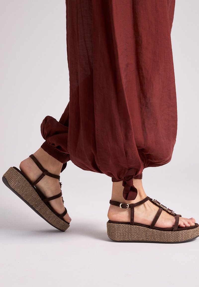 Pies con sandalias de plataforma marrones adornadas con detalles florales, combinadas con pantalones sueltos de color óxido con dobladillo anudado, sobre un fondo claro.