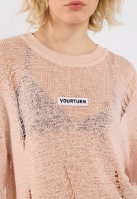 Jasnoróżowy sweter dziany o luźnym kroju, z przetarciami i czarną metką "YOURTURN" z przodu. Teksturowany, przewiewny materiał.