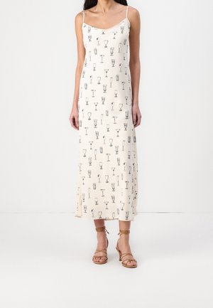 Femme portant une robe maxi crème sans manches avec des imprimés de verres à cocktail noirs et des sandales à talons hauts tan à brides.