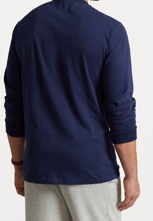 Homme portant une chemise à manches longues bleu marine retroussées et un pantalon gris clair, vu de dos sur un fond uni.