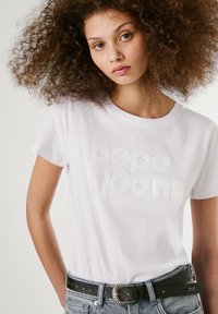 Hvit bomulls t-skjorte med hevet "pepe jeans" tekst. Kombinert med et svart belte med en sirkulær metallspenne, og blå denimshorts.