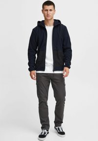 Sudadera con cremallera en color azul marino con bolsillos texturizados negros, combinada con una camiseta blanca y pantalones cargo oscuros. Zapatillas negras con detalles en blanco.