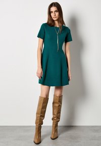 Abito corto teal a maniche corte con orlo svasato, realizzato in tessuto testurizzato, abbinato a stivali al ginocchio in pelle marrone. Scollo semplice e linee di design.