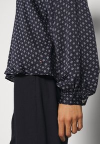 Blusa navy con motivo geometrico bianco, vestibilità ampia, maniche a sbuffo e polsini con bottoni. Presenta un piccolo logo vicino all'orlo.