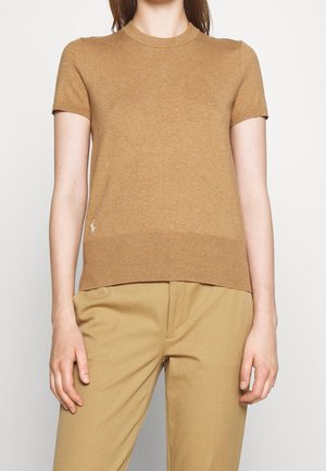 Femme portant un pull en maille à manches courtes couleur camel et un pantalon beige assorti, debout devant un fond clair uni.