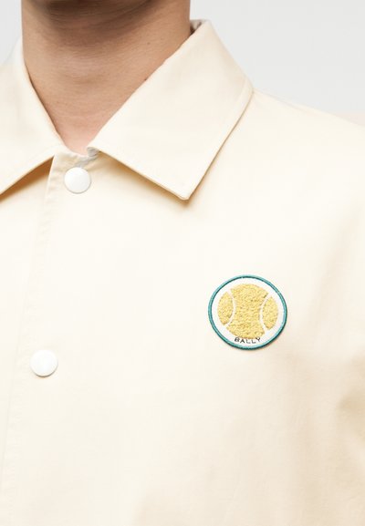 Κοντινό πλάνο μπεζ μπουφάν με λευκά κουμπιά snap και κεντημένο έμβλημα με κίτρινη μπάλα τένις περιγραμμένη με τιρκουάζ στο στήθος.