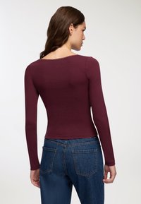 Bordowy top z długim rękawem i okrągłym dekoltem, ozdobiony przeszyciami wzdłuż krawędzi, zestawiony z niebieskimi spodniami z wysokim stanem.