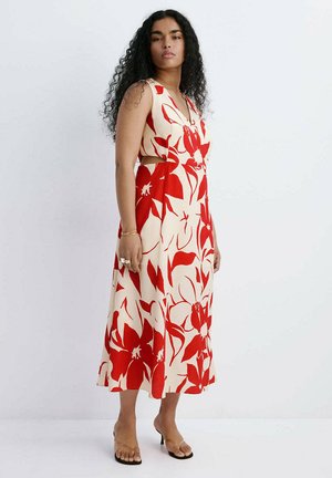 Femme aux longs cheveux bouclés portant une robe crème sans manches avec de grandes impressions florales rouges et des sandales à talons noires.