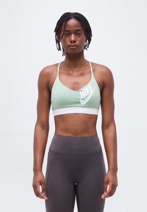 Atletische vrouw met korte dreadlocks, gekleed in een lichtgroene Nike sportbeha en hooggetailleerde donkergrijze legging, staand tegen een effen achtergrond.
