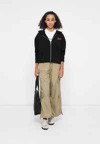 Veste à zip noir avec logo rose, pantalon cargo beige à jambes larges, et baskets blanches. La tenue comprend un tote bag noir.