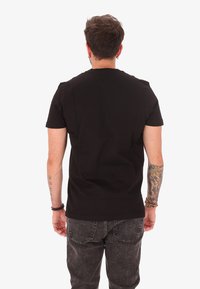 T-shirt nera in cotone con maniche corte, scollatura rotonda e silhouette aderente, mostrata di schiena. Tatuaggio visibile sul braccio sinistro.