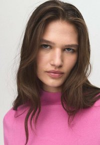 Ein Model mit langen, braunen Haaren trägt einen taillierten, leuchtend pinken Pullover mit hohem Kragen. Glatte Haut und Make-up in neutralen Tönen.