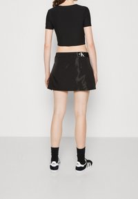 Un top court noir et une jupe A-line noire et brillante avec une ceinture CK. Le mannequin porte des chaussettes noires et des baskets Adidas avec des accents blancs.