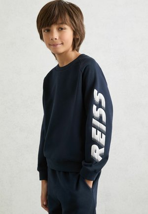 Tamsiai mėlynas džemperis iš minkštos medžiagos, su apvalia iškirpte ir baltais grafiškai užrašytais užrašais kairėje rankovėje. Sukurtas laisvo kirpimo.