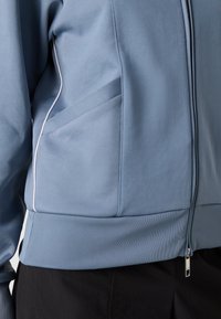 Light blue zip-up jacket with ribbed hem, featuring contrast stitching and angled side pockets. Smooth fabric with a soft texture.