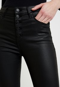 Černé džíny s vysokým pasem, které mají hladkou texturu, zapínání na pět knoflíků a boční kapsy pro větší funkčnost.