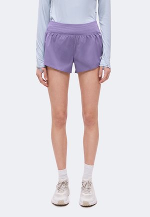 Person iført lyse lilla træningsshorts, hvide sneakers, hvide sokker og en lyseblå langærmet top synlig fra nakke til fødder.