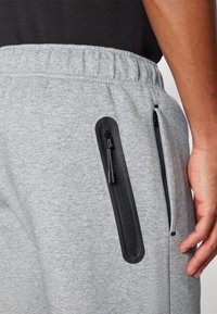 Primo piano di una persona che indossa pantaloni della tuta grigi con due tasche verticali nere con zip ai lati e una maglietta nera.