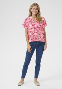 Blusa rosa a maniche corte con un motivo floreale, abbinata a jeans skinny blu scuro e ballerine di colore chiaro. Tessuto morbido con una vestibilità comoda.