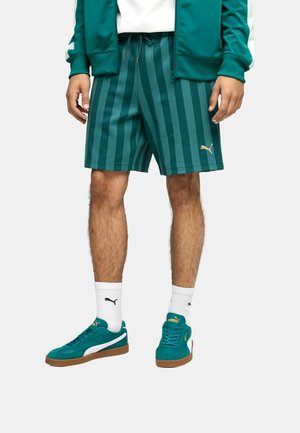 Mężczyzna ubrany w turkusowe szorty w paski, pasujące turkusowe trampki z białym logo Puma, białe skarpetki Puma oraz zieloną kurtkę z białymi akcentami.