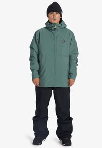 Zelená nepremokavá bunda s predným zipsom, nastaviteľnou kapucňou a nášivkou loga. V kombinácii s voľnými čiernymi nohavicami a čiernymi čižmami.
