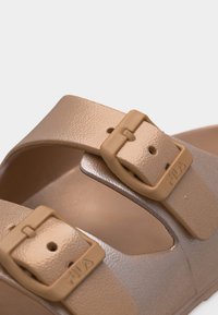 Κοντινή λήψη από μπεζ λουράκια σανδαλιών με ρυθμιζόμενες αγκράφες που φέρουν ανάγλυφο το λογότυπο "FILA" σε ανοιχτό φόντο.