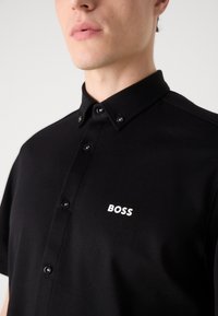 Juodas trumpas rankovėmis marškiniai su knopkiniu apykakle, su baltu "BOSS" logotipu ant krūtinės ir juodais knopkių akcentais. Glotni tekstūra.