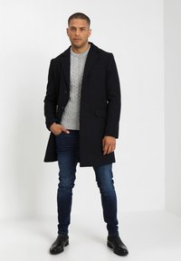 Abrigo de lana azul marino con un corte entallado, grandes botones y bolsillos laterales, combinado con un suéter de punto trenzado gris y jeans de mezclilla oscura. Botines negros.