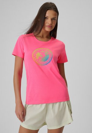 Kobieta z długimi brązowymi włosami, ubrana w jasnoróżowy T-shirt z kolorową, okrągłą grafiką oraz jasnobeżowe szorty na szarym tle.
