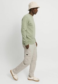 Lysegrønn genser, beige cargobukser med lommer, kremfargede joggesko og en beige bøttehatt. Enkelt, avslappet passform og komfortabelt design.