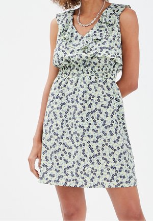 Femme portant une robe sans manches longueur genou avec un petit imprimé floral bleu et un col à volants, accessoirisée d'un collier chaîne en argent.