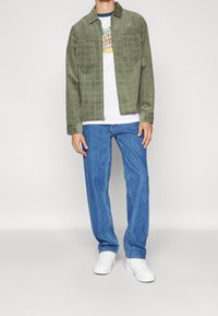 Olijfgroene corduroy jas met voorzakken, gecombineerd met een witte graphic t-shirt en blauwe losvallende jeans. Witte sneakers maken de look compleet.