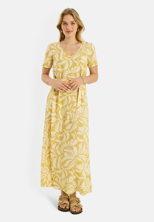 Mujer con vestido maxi de escote en V con estampado floral amarillo y blanco, mangas cortas y sandalias beige, de pie frente a un fondo liso.