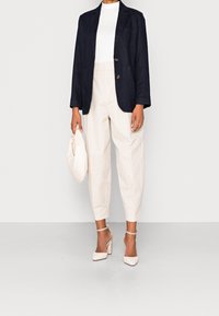 Navy linen blazer with two buttons, white fitted top, beige pleated trousers, and light beige block-heel shoes, complemented by a textured clutch.