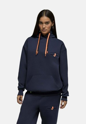 Femme portant un sweat à capuche bleu marine et un pantalon assorti avec des cordons orange et de petits détails orange sur les poches, debout devant un fond uni.