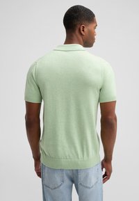 Koszulka polo z krótkim rękawem w jasnej zielonej dzianinie. Posiada ściągaczowy kołnierzyk i dół. Noszona z jasnymi dżinsami, z widokiem z tyłu.